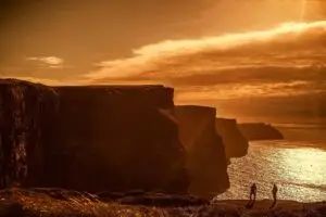 Two people walking near the dramatic and serene Cliffs of Moher at sunset, highlighting the natural beauty and tourist appeal of these famous Irish seaside cliffs.