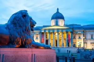 Twilight view of the National Gallery and a lion statue at Trafalgar Square in London, England, showcasing historic architecture and art culture.