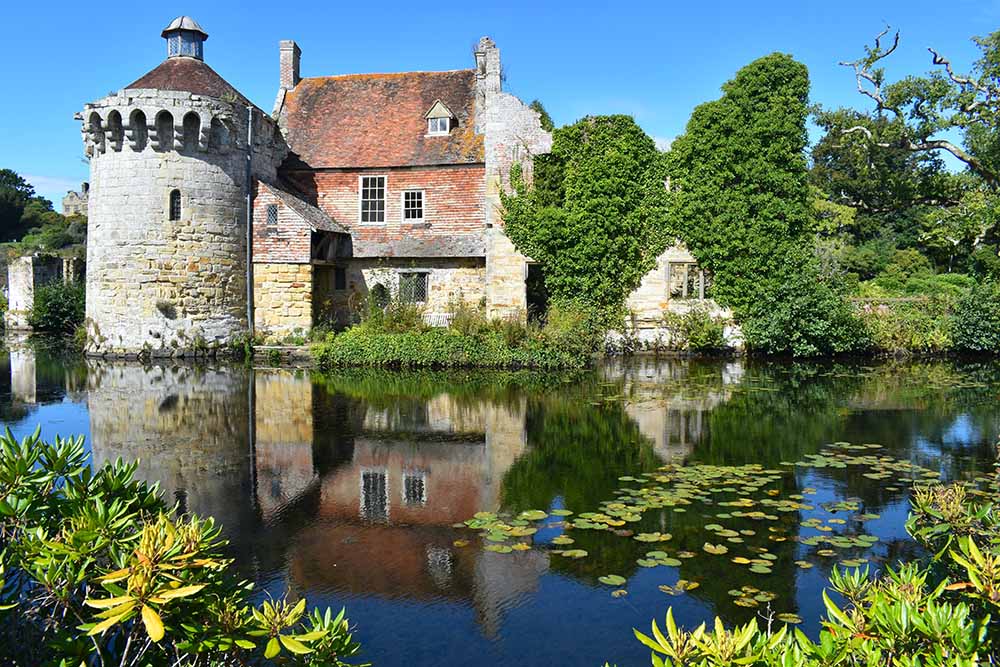 Scotney Castle reflected in a serene pond surrounded by lush greenery under a clear blue sky in England, showcasing picturesque medieval architecture.