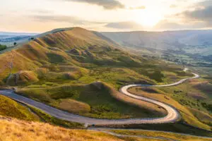 Scenic view of a winding road through the rolling hills of the Peak District at sunset, highlighting the golden light and lush landscapes ideal for exploring UK nature and travel destinations.