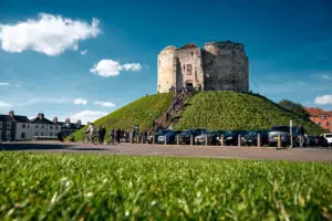Visitors exploring the historic Clifford's Tower on a sunny day in York, United Kingdom, surrounded by lush greenery and clear blue skies.
