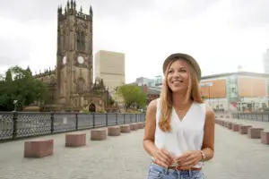 Young woman in a white blouse and hat smiling near Manchester Cathedral, showcasing urban tourism in Manchester, UK.